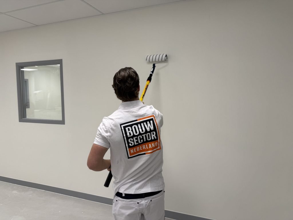 Schilder van Bouwsector Nederland rolt een lichte wand met een verlengsteel, naast een binnenraam